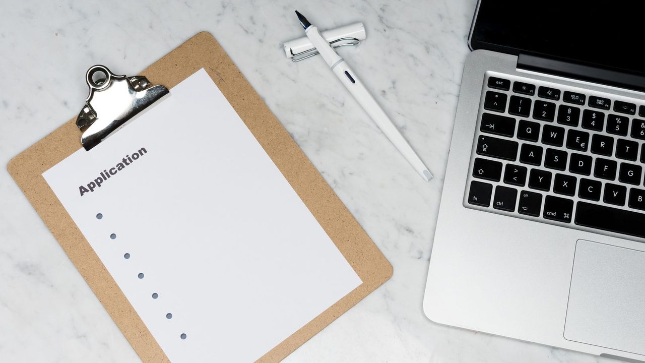Clipboard with a blank application form, a pen and a laptop on a marble desk, no people or logos